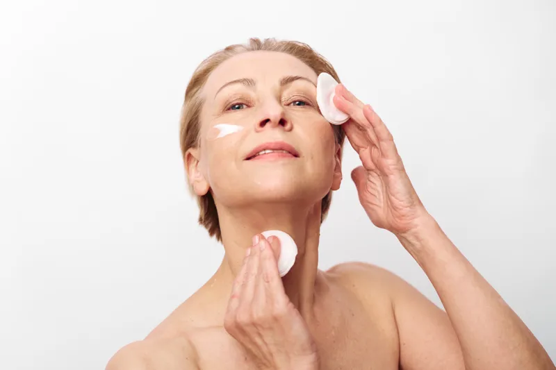 Mature woman applying facial cream with cotton pads on clean skin, demonstrating skincare routine and self-care for healthy aging and natural beauty on white background.