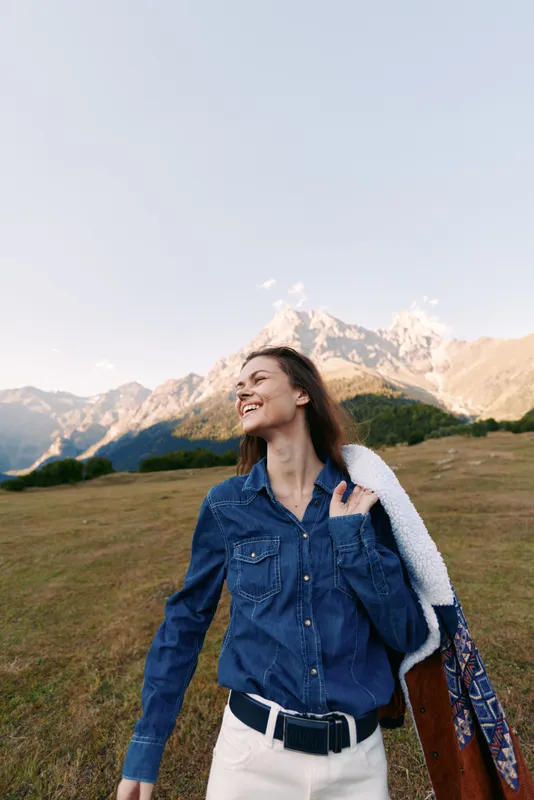 Woman smiling in denim shirt and jacket over shoulder in mountain meadow outdoors, happy portrait of travel lifestyle, enjoying nature and fresh air with scenic peaks behind.