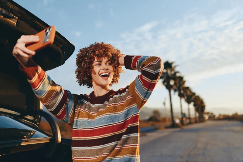 Woman lifestyle, curly hair smiling, stands by car trunk, holds smartphone near open door, wearing casual striped sweater, road and trees in distance, sunset outdoor travel atmosphere