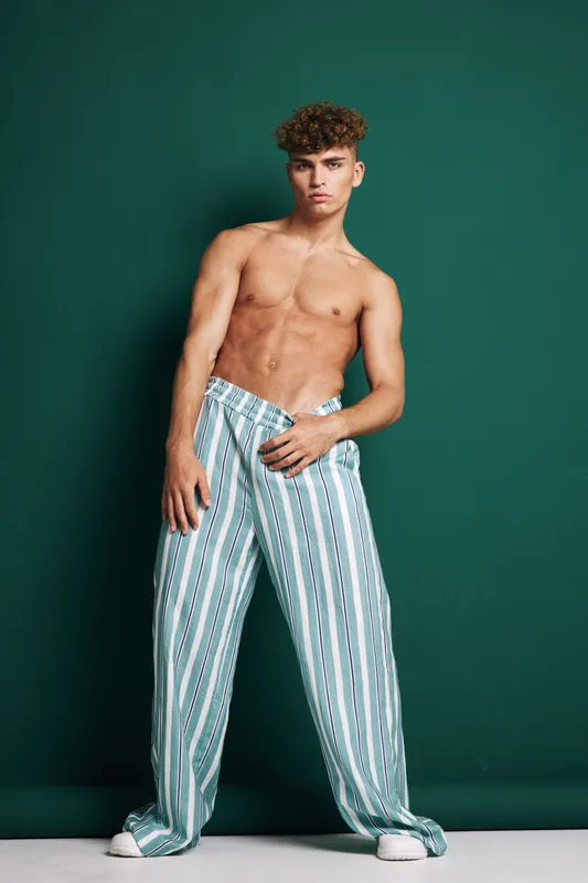 Young man with curly hair posing shirtless in striped wide pants against green background, fashion model with confident expression and casual style in studio.