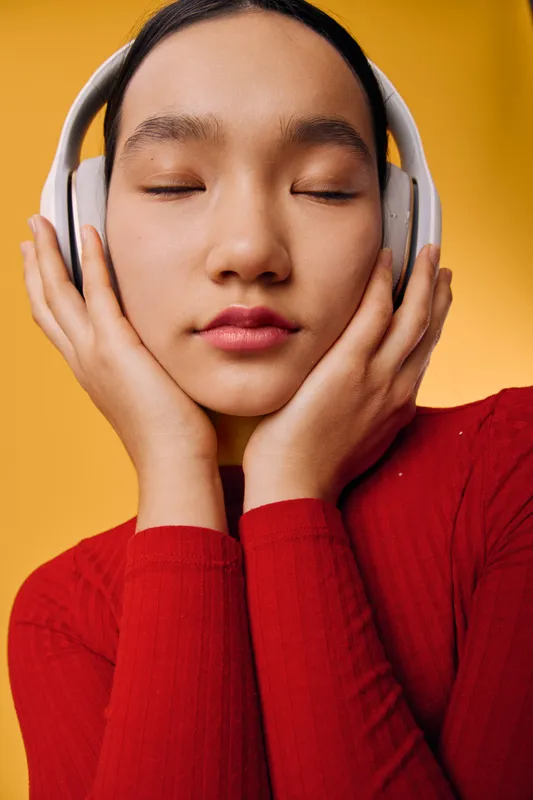Young woman with closed eyes wearing headphones and red sweater feeling calm and relaxed while listening to music on vibrant yellow background. Closeup portrait with peaceful expression.