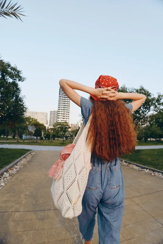 Lifestyle woman in modern boho-western denim street style with long red hair walking outdoors under blue sky, carrying a large crochet bag in warm film color tones.