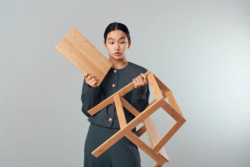 Young woman in formal outfit holding a wooden chair and plank looking surprised with gray background. Studio portrait of unboxing, furniture, product, wooden, unpacking and casual style concept.