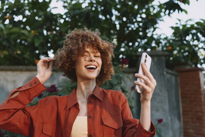 A cheerful woman with curly hair in an orange jacket takes a selfie in a sunny outdoor setting, capturing a candid lifestyle moment. Focus on joy, self-expression, and everyday positivity