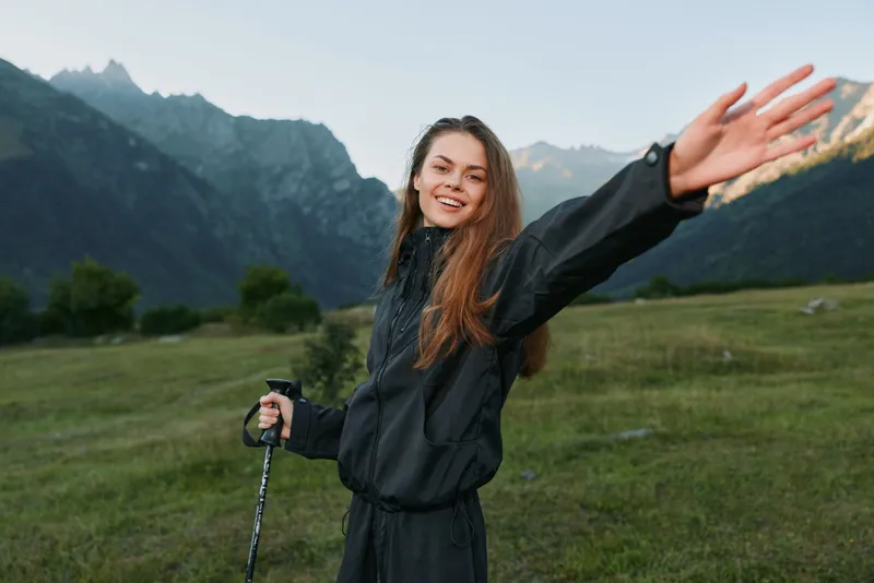 A cheerful hiker stands in a mountain valley, smiling with arms raised, wearing a dark jacket and trekking pole, enjoying the outdoor landscape and fresh air.