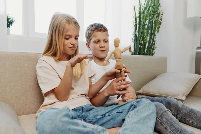 Two children sitting on a sofa play with a wooden mannequin toy in a bright living room, showing creativity and sibling bonding with casual clothing and natural light.