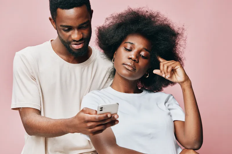 Young African couple enjoying a moment while using a smartphone, smiling and interacting in a cozy pink background A modern portrayal of love and connection