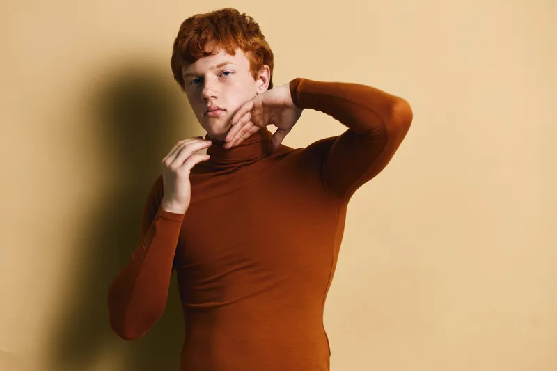 Young man with red hair posing in brown turtleneck sweater against beige background, fashion portrait with thoughtful expression and casual style for model shoot.