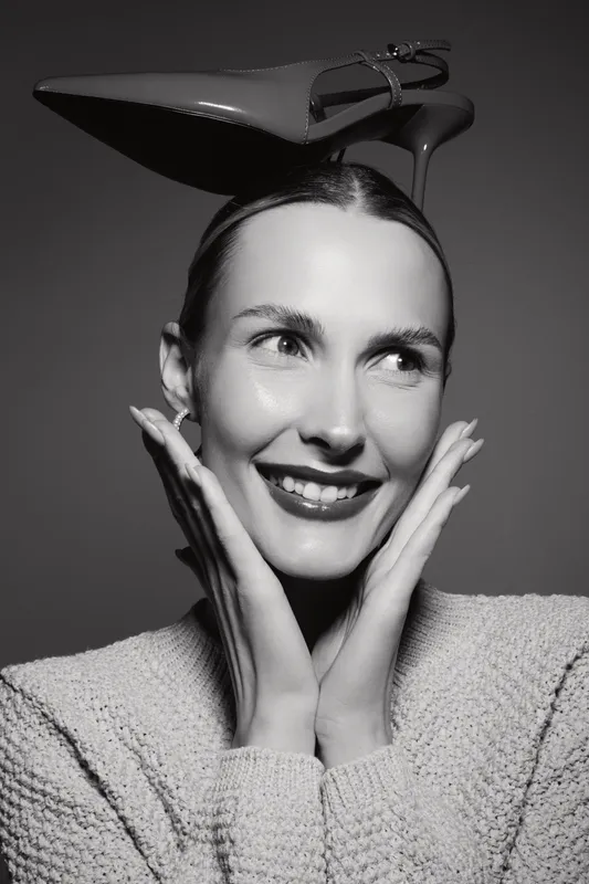 Young white woman with light skin smiles joyfully holding her face with both hands, balancing high heel shoe on her head. Concept of playful fashion and beauty portrait.
