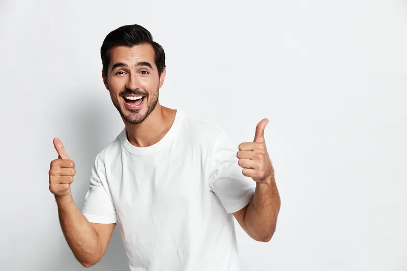 Happy smiling young caucasian man giving thumbs up and looking at the camera, wearing a plain white t-shirt, isolated on a light neutral background. People lifestyle concept.