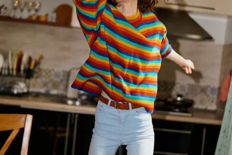 Joyful person in a striped colorful sweater, indoors at home, mid movement, natural light, warm tones, casual lifestyle, kitchen backdrop, candid moment with soft breeze, relaxed posture