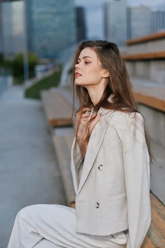 Woman in beige suit sitting on urban bench with pensive expression, natural makeup, and modern office background, stylish fashion portrait outdoors