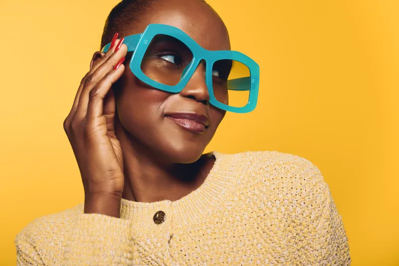 Portrait of confident African woman with dark skin wearing large blue sunglasses and beige sweater on yellow background. Concept of fashion, style, and beauty.