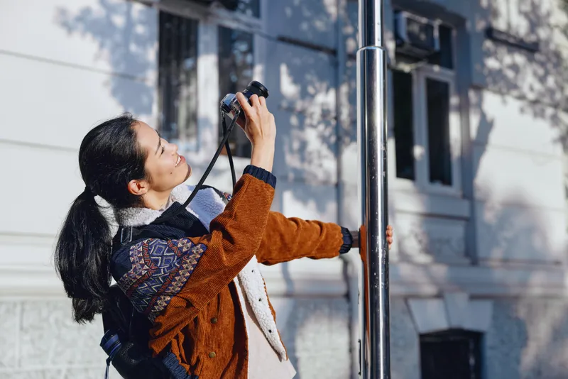 Woman photographer on a street corner, camera raised, scarf fluttering and jacket warm, daylight highlighting urban outdoors and candid moment, capturing a dynamic shot and textured scene
