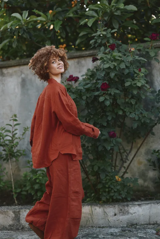 Young woman with curly hair walks along a garden path in a rust orange jumpsuit, smiling softly as blooming roses frame the scene