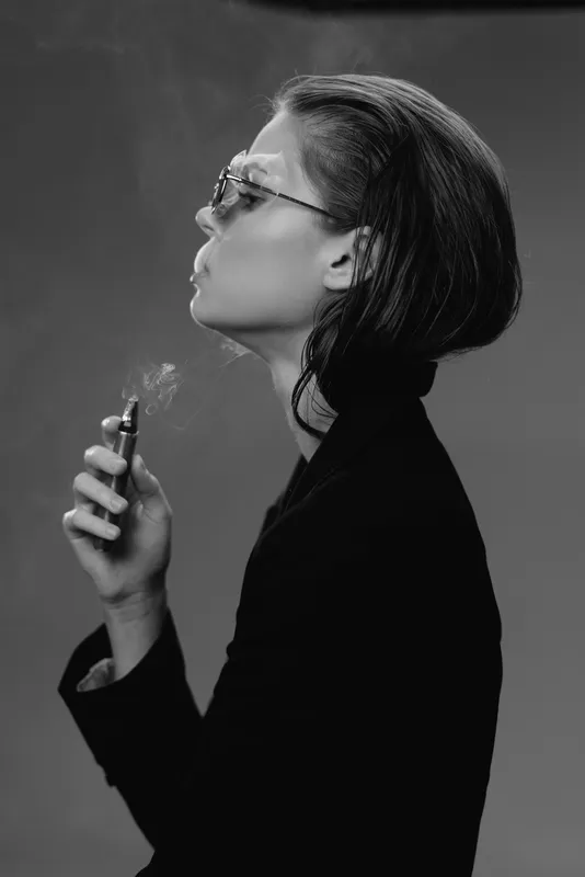 Black and white profile of a stylish woman smoking vape with sunglasses and short hair in dark clothing