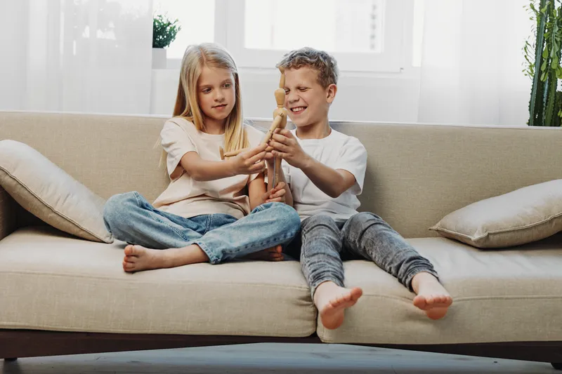 Two children sitting on a beige sofa playing with a wooden toy, enjoying a relaxed moment in a bright living room. Concept of childhood, play, and sibling bonding.
