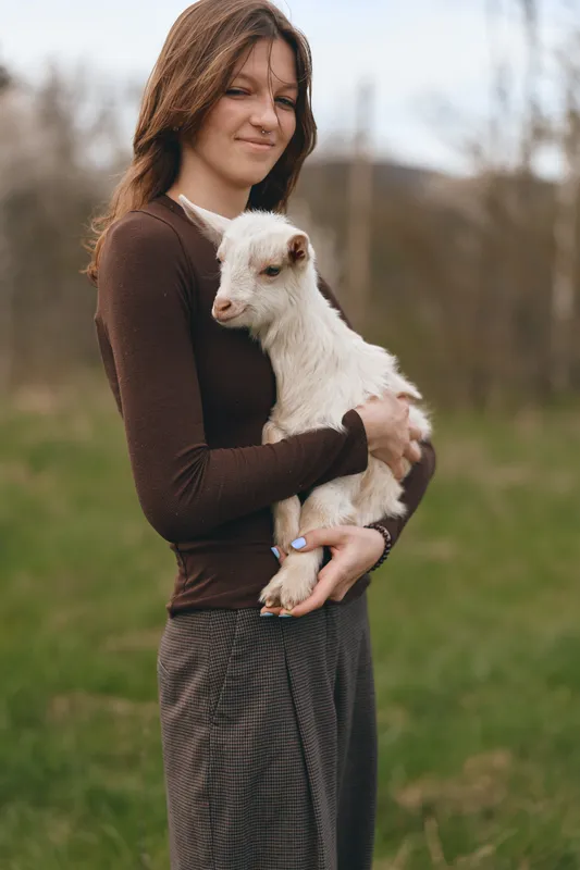 A woman tenderly holds a goat kid in a grassy field, capturing rural life and gentle livestock bonding in a calm outdoor moment, highlighting care, warmth, and connection.