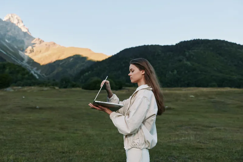 A focused female professional stands in a wide open field with a laptop and stylus, working outdoors among mountains, conveying innovation and remote productivity.