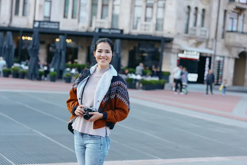 Photography of a smile on a street with a camera, showcasing urban fashion in a warm jacket, outdoors during autumn, capturing candid lifestyle moments and cityscape ambiance for street photography.