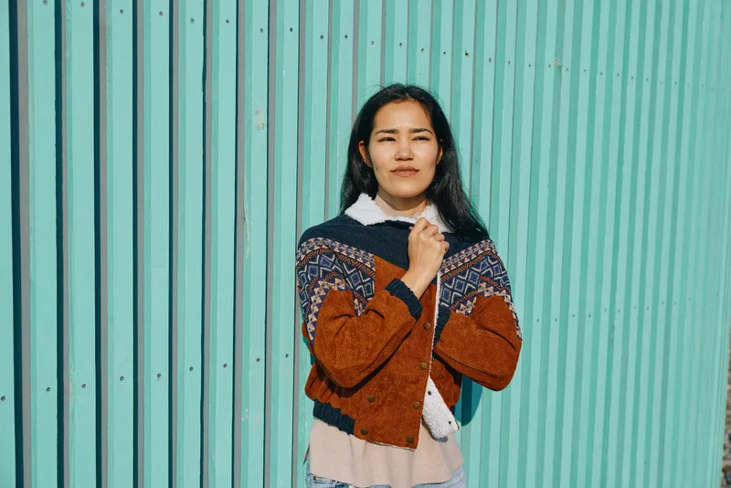 Female in knitted cardigan and warm sweater stands before a teal wall, showcasing casual fashion and cozy texture with a confident, relaxed outdoor pose in a vibrant urban setting.