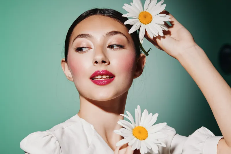 Portrait of a creative young woman holding daisies near her face with glossy lips and flawless skin on a mint background. Concept for beauty fashion and natural elegance.