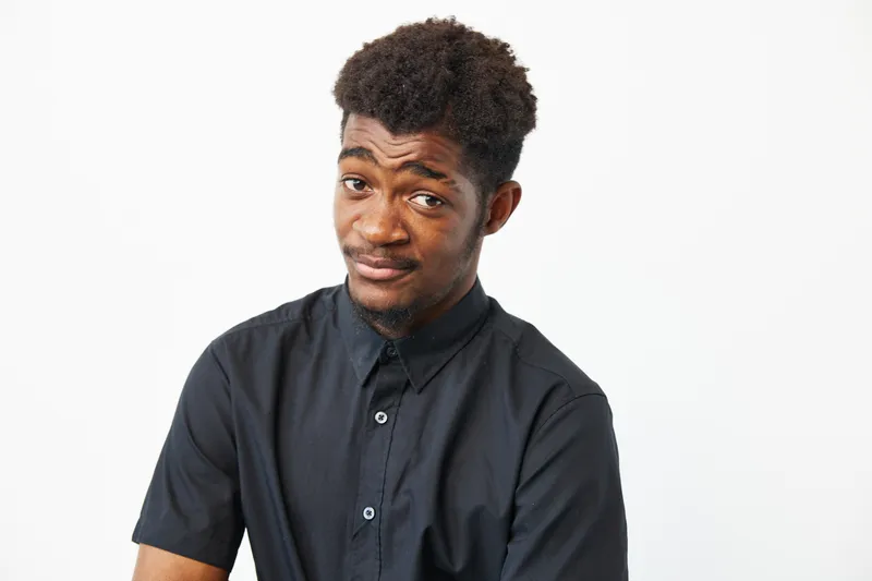 Black man with a playful expression, wearing a stylish black shirt, showcasing confidence and charm against a clean white background.