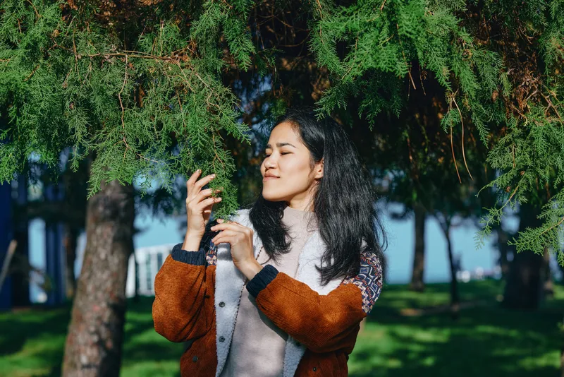 Nature scene with a woman in a warm sweater enjoying sunlight among trees, admiring a leaf close up in a peaceful park setting, conveying calm, curiosity and natural harmony