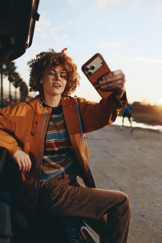 Curly hair woman in casual jacket sits outdoors at sunset, holding a smartphone for a selfie portrait. Warm golden light and lifestyle mood, urban road background, relaxed travel pose.