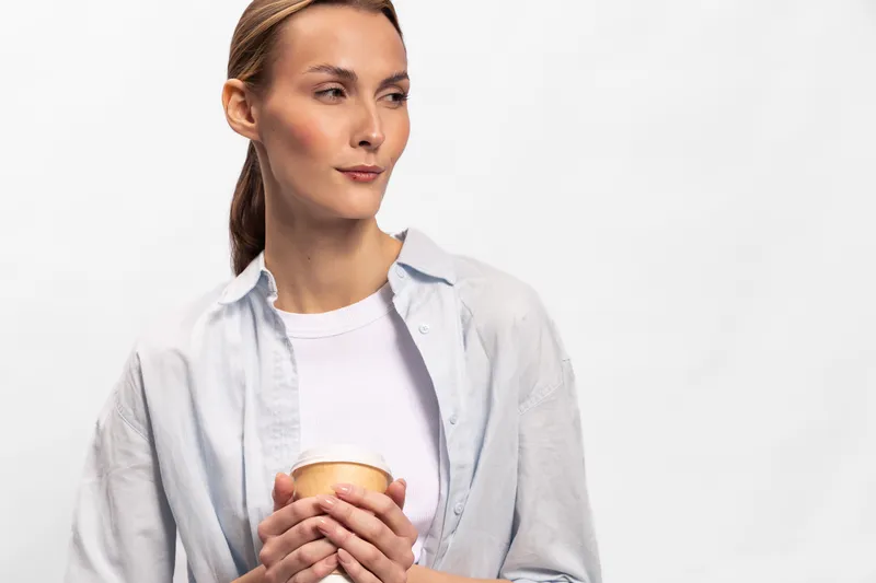 Calm young woman with light skin and tied hair holds a cup wearing casual shirt and white top, looking away thoughtfully against a plain white background. Concept of relaxation and contemplation.