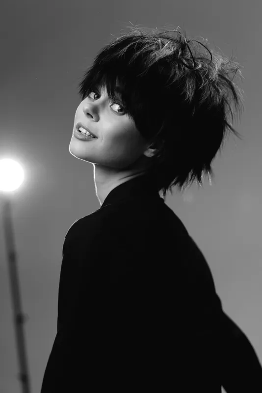 Black and white portrait of a young woman with tousled hair looking over her shoulder, showing a thoughtful expression with soft lighting.