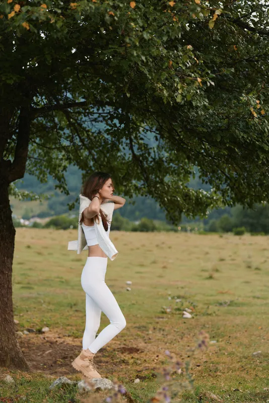 Young woman in white outfit stands under a large tree in a grassy field adjusting her hair with a calm and thoughtful expression during a peaceful outdoor moment.