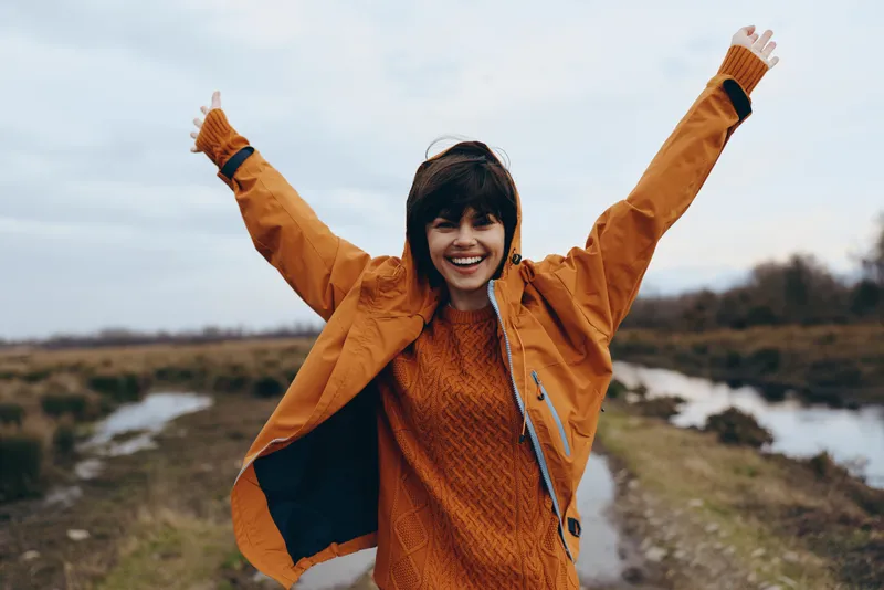 Woman lifestyle outdoors smiling autumn jacket scarf nature happiness, arms raised in celebration, warm orange knit clothing, countryside path, soft wind, clear sky, joyful expression