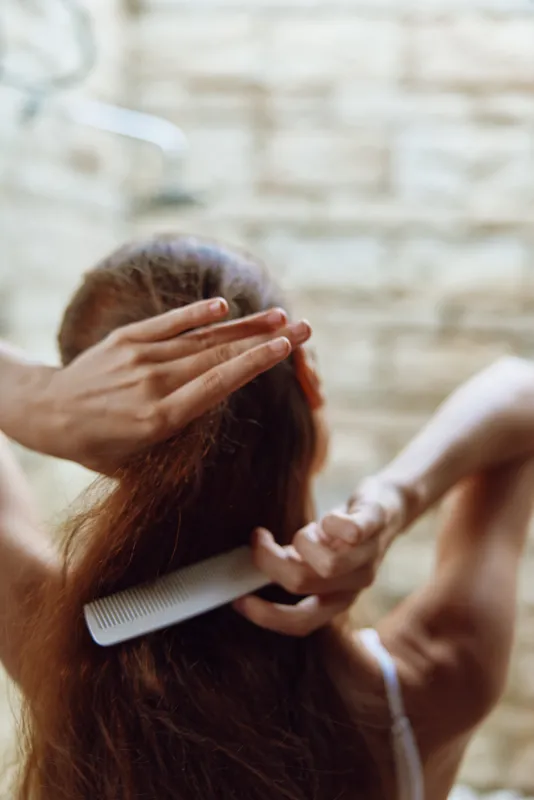 Woman combing long brown hair in a bright bathroom with brick wall, natural light highlighting healthy shiny strands and gentle morning self-care routine.