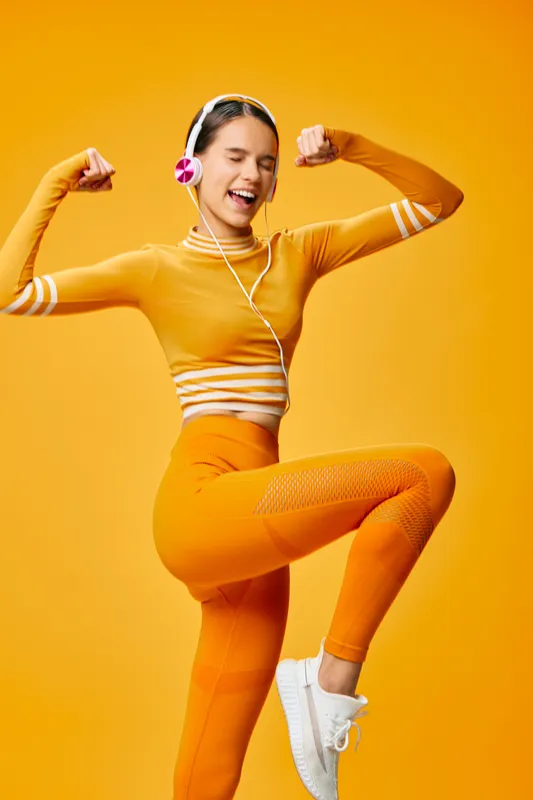 Woman fitness enthusiast in bright orange athletic wear, headphones on, performing a dynamic dance-like workout against a vivid orange backdrop.
