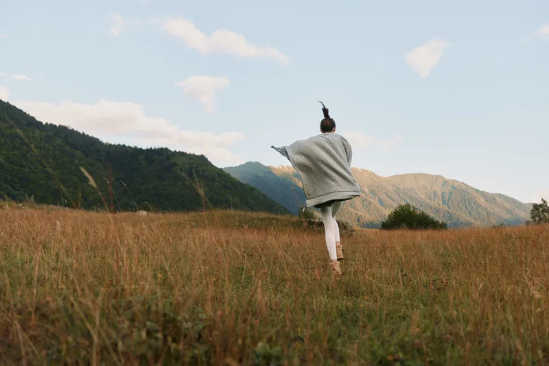 Woman in a light sweater runs through a tall grass field toward distant mountains, capturing a carefree outdoor lifestyle and fresh air.