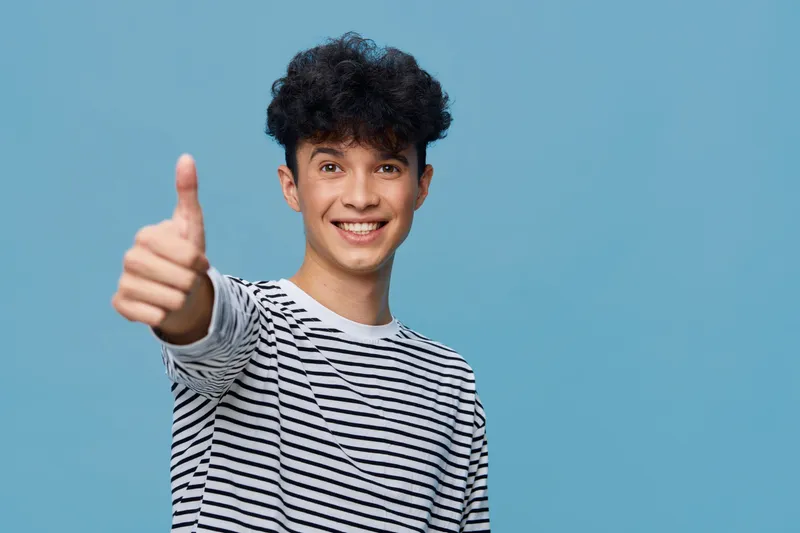 smiling young man in striped shirt giving thumbs up gesture against blue background with curly hair and cheerful expression