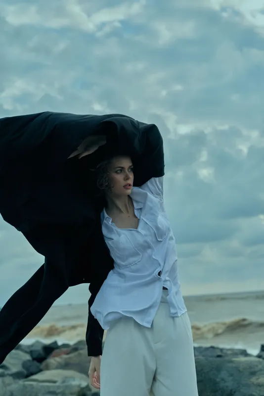 Windblown fashion on a beach shoots a model in white shirt and light trousers beneath a flowing cloak, ocean horizon behind, creating movement and dramatic mood.
