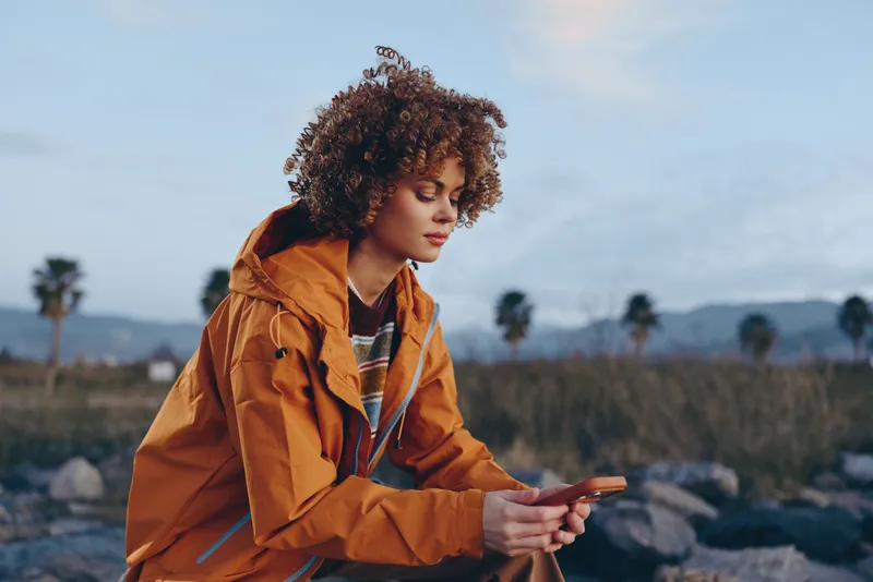 Woman lifestyle outdoors, curly haired woman in orange jacket, standing among rocks at sunset, holding smartphone, relaxed gaze, soft breeze, natural tones, wide rural background