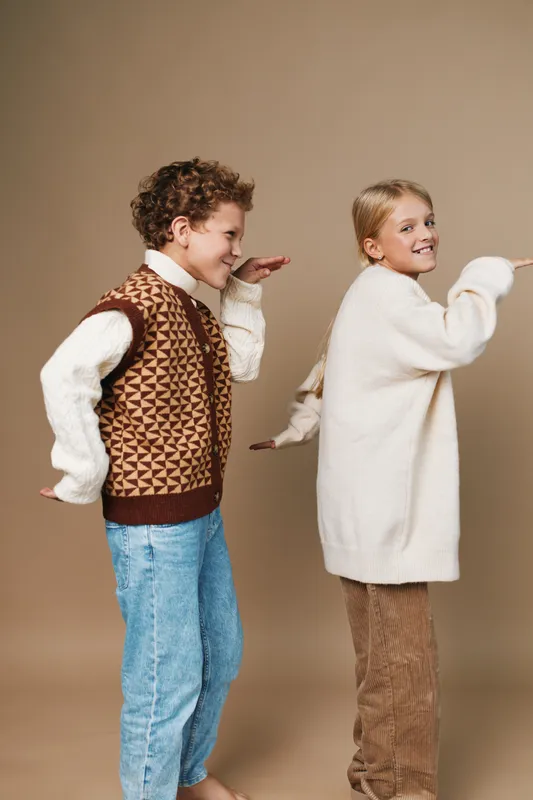 Two children pose playfully in a studio, back to back with arms extended as if flying, wearing cozy sweaters and jeans, warm beige backdrop, lighthearted and lively.