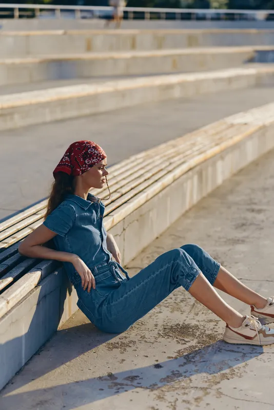 Young woman relaxing outdoors on concrete steps wearing casual blue outfit and red headscarf in warm sunlight with a peaceful expression.