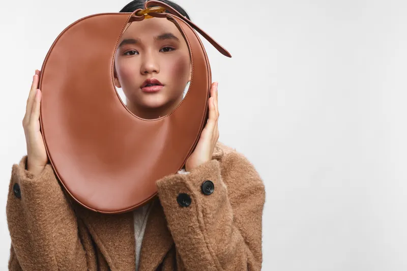Asian woman wears a cozy coat and holds a round leather bag close to her face, creating an avant garde fashion portrait with soft studio lighting and a bold yet intimate atmosphere.