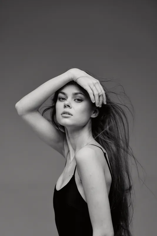 Black and white portrait of a young woman with long hair posing elegantly in a simple black top, showcasing beauty and style.