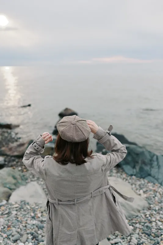 Woman in beige coat and hat stands on rocky shore, looking at calm sea under cloudy sky. Concept of solitude, contemplation, and peaceful nature moment by the ocean.