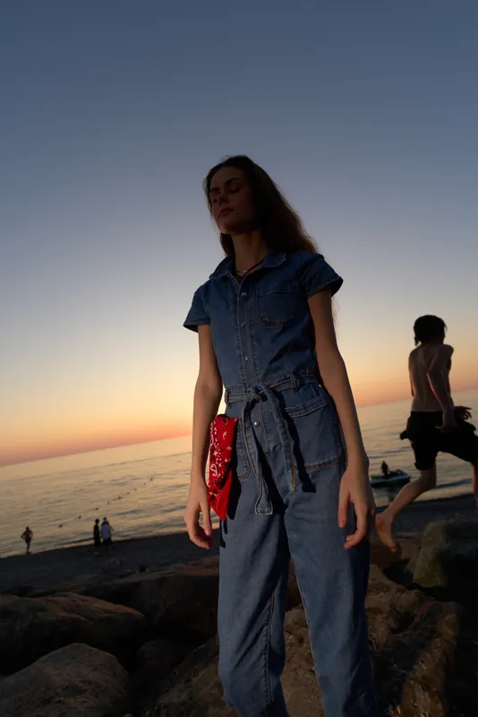 lifestyle woman in modern boho-western denim street style standing by the beach at sunset, with film color tones creating a warm atmospheric mood