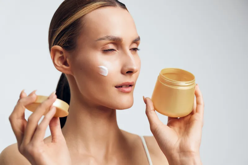 Young woman applying cream with a cosmetic jar on her cheek, close-up on a light background, conveying a calm skincare moment and self care routine, soft focus, warm mood.