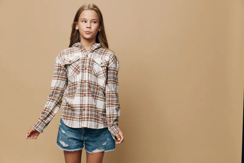 Girl in plaid shirt and denim shorts standing with relaxed pose on beige background. Young female child with long hair wearing casual summer outfit for fashion.