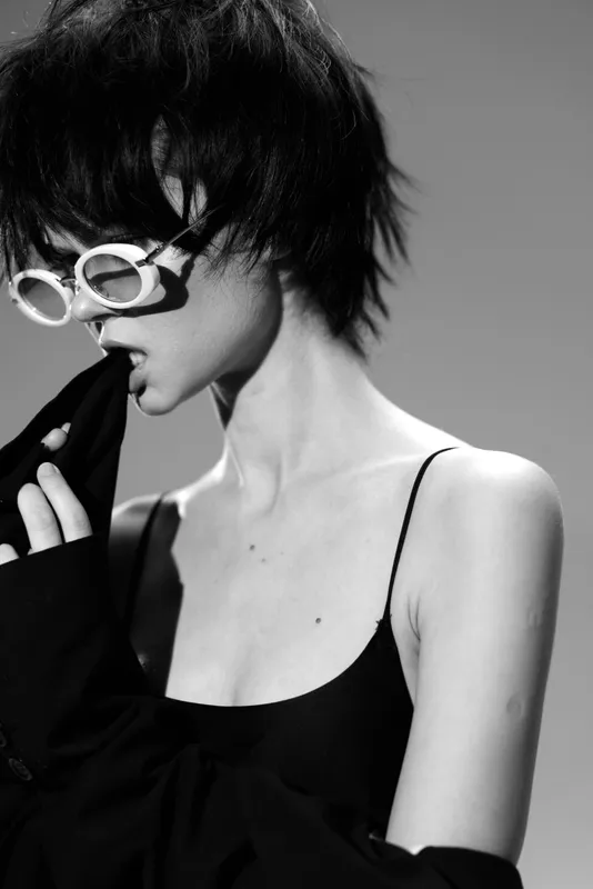 Black and white portrait of a stylish woman wearing white sunglasses and a black top, expressing a bold and edgy mood.