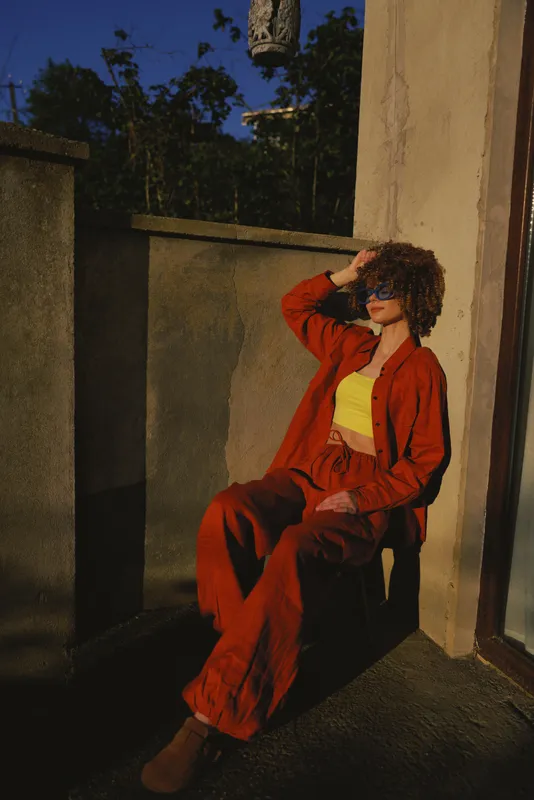 Casual lifestyle photograph of a woman sitting on a balcony, wearing an orange suit with a yellow top, posing thoughtfully under warm sunlight in an urban outdoor setting