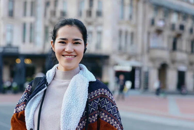 Woman with a warm smile outdoors on a city street, layered coat and scarf, casual fashion, daylight in an urban setting for lifestyle, travel and street style use